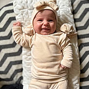 Elley a rejoint le concours — aidez-le/la à gagner de superbes lots ! baby, blanket, cheerful, clothing, cozy, face, hand, hat, headband, indoor, infant, leg, lying_down, onesie, pillow, plush, portrait, smile, soft_texture, zigzag_pattern