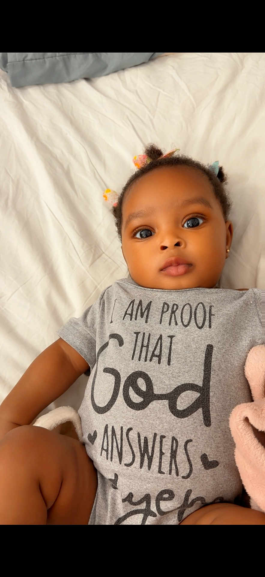 Giovanna joined the competition — help win amazing prizes! baby, infant, child, onesie, text_on_clothing, plush_toy, bed, white_bed_sheet, hair_ties, cute, portrait, indoor, skin, face, eyes, mouth, hand, lying_down, clothing, soft_toy