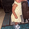 Cimani is registered to the contest to win money with this photo: toddler, child, yellow_dress, sneakers, living_room, couch, rug, curly_hair, standing, indoor, furniture, carpet, person, cute, expression, small_child, home, floor, casual_clothing, young