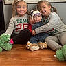 animal_slippers, baby, casual_clothing, child, cozy, family, floor, gray_wall, happy, helmet, hugging, indoor, long_hair, overalls, pigtails, sitting, slippers, smiling, toddler, wooden_floor