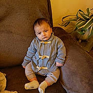 Léo participe au concours pour gagner de l'argent avec cette photo : baby, infant, child, sitting, couch, sofa, brown_couch, sweater, knitwear, socks, blanket, potted_plant, houseplant, living_room, indoor, portrait, expression, frown, cushion, furniture