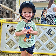 Leo participe au concours pour gagner de l'argent avec cette photo : child, boy, helmet, green_tshirt, paw_patrol, shorts, fence, hay, outdoor, smile, person, tree, women, background, building, daylight, casual_clothing, playful, happy, standing