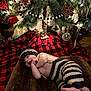 baby, infant, christmas_tree, ornaments, blanket, knit_clothing, hat, striped_pants, decorations, lights, plaid, groot_ornament, pine_cones, red_rose, toddler, holiday, festive, indoor, smiling, cute