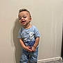 toddler, child, boy, curly_hair, standing, blue_tshirt, jeans, socks, carpet, wall, happy, expression, indoor, person, clothing, smiling, feet, young_child, casual, portrait
