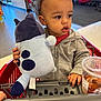 toddler, child, shopping_cart, plush_toy, curly_hair, grey_jacket, indoor, store, drink_cup, red_cart, hand, casual_clothing, young_child, shopping, candid, gaze, cute, retail, portrait, curiosity