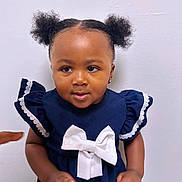 Nefertiti is registered to the contest to win money with this photo: toddler, child, girl, navy_dress, white_bow, curly_hair, puffs, earrings, standing, indoor, white_wall, cute, portrait, skin, hand, pointing, smiling, short_sleeves, lace_trim, baby