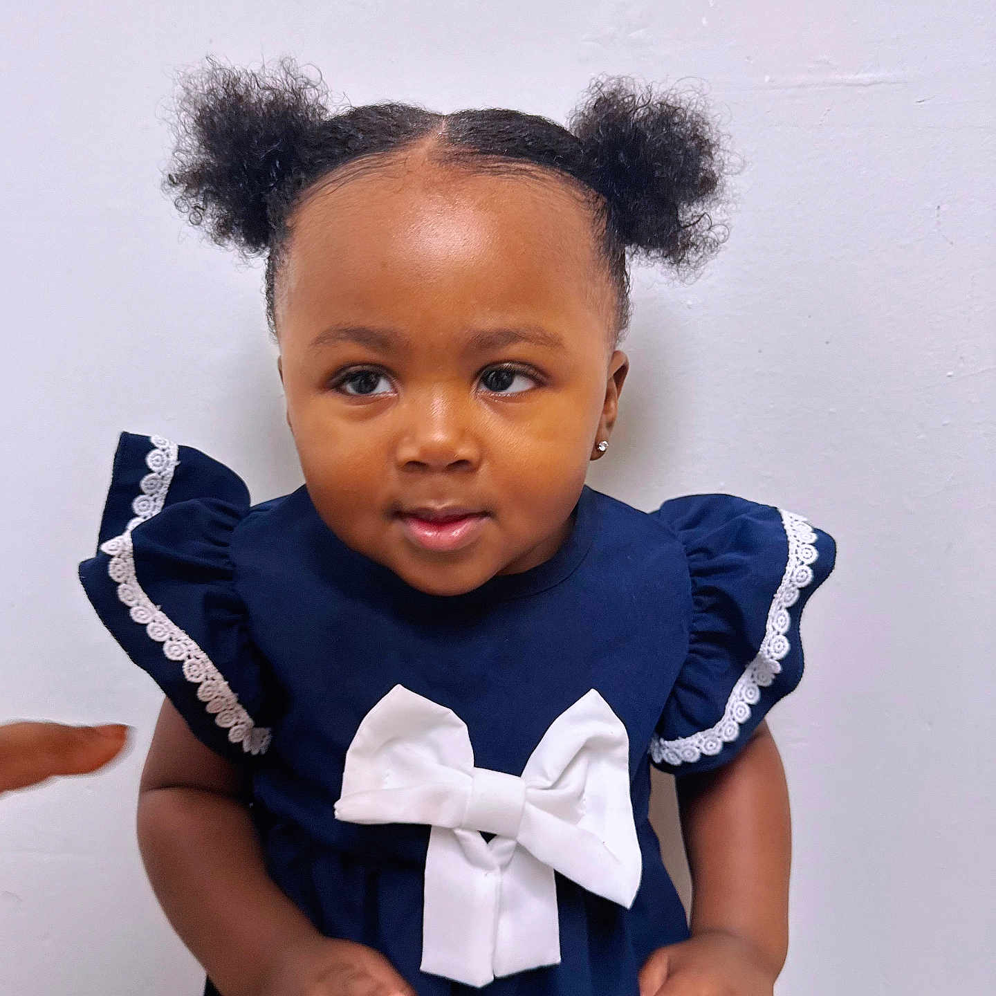 Nefertiti is registered to the contest to win money with this photo: baby, child, curly_hair, cute, earrings, girl, hand, indoor, lace_trim, navy_dress, pointing, portrait, puffs, short_sleeves, skin, smiling, standing, toddler, white_bow, white_wall