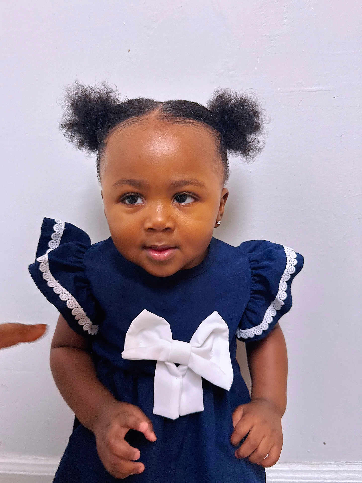 Nefertiti is registered to the contest to win money with this photo: toddler, child, girl, navy_dress, white_bow, curly_hair, puffs, earrings, standing, indoor, white_wall, cute, portrait, skin, hand, pointing, smiling, short_sleeves, lace_trim, baby