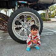 Cyphyr Pineda joined the competition — help win amazing prizes! accessory, asphalt, blue_crocs, boy, casual, child, clothing, cute, daylight, happy, outdoor, parking_lot, playful, smiling, squatting, tire, toy, truck, vehicle, wheel