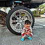 accessory, asphalt, blue_crocs, boy, casual, child, clothing, cute, daylight, happy, outdoor, parking_lot, playful, smiling, squatting, tire, toy, truck, vehicle, wheel