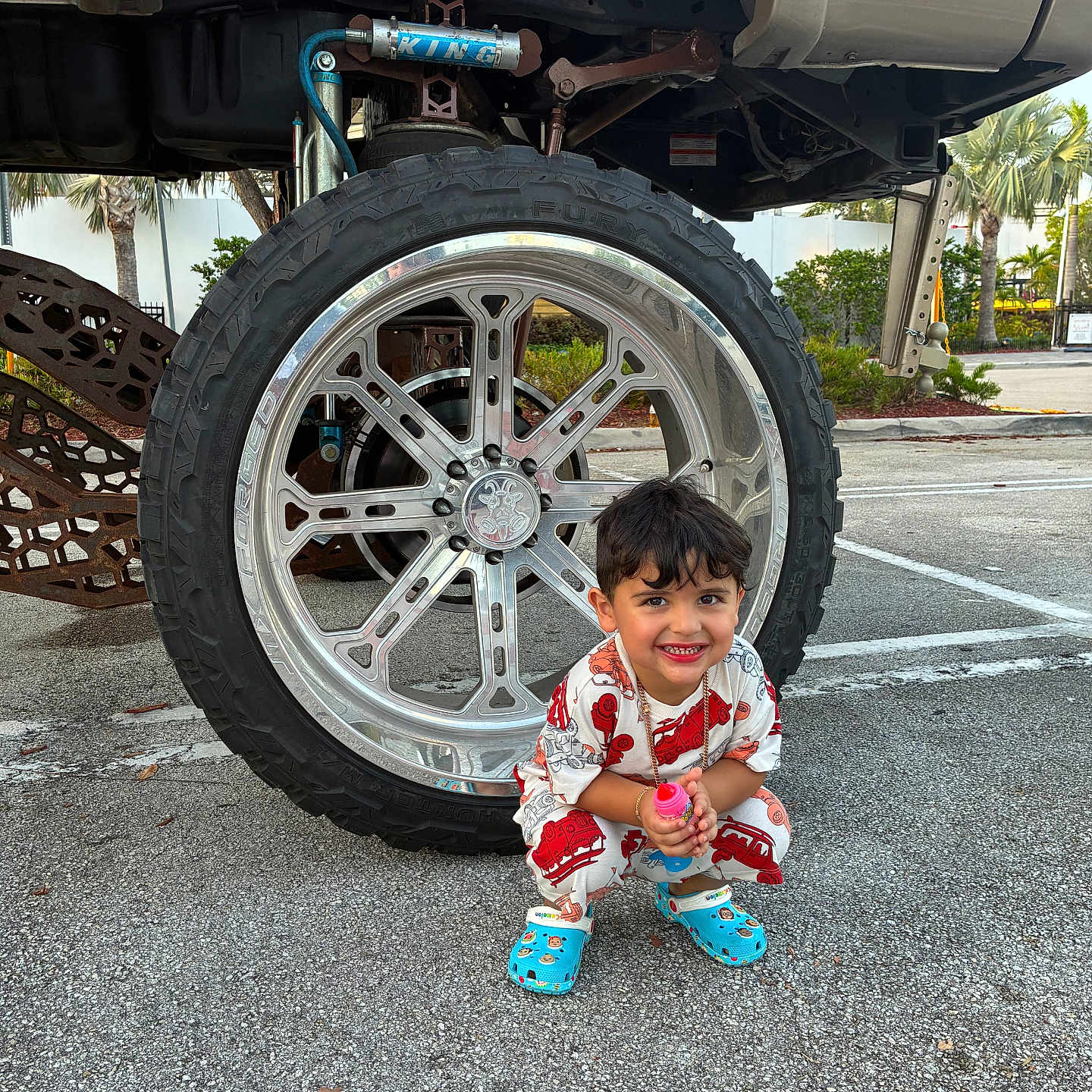 Cyphyr Pineda joined the competition — help win amazing prizes! accessory, asphalt, blue_crocs, boy, casual, child, clothing, cute, daylight, happy, outdoor, parking_lot, playful, smiling, squatting, tire, toy, truck, vehicle, wheel