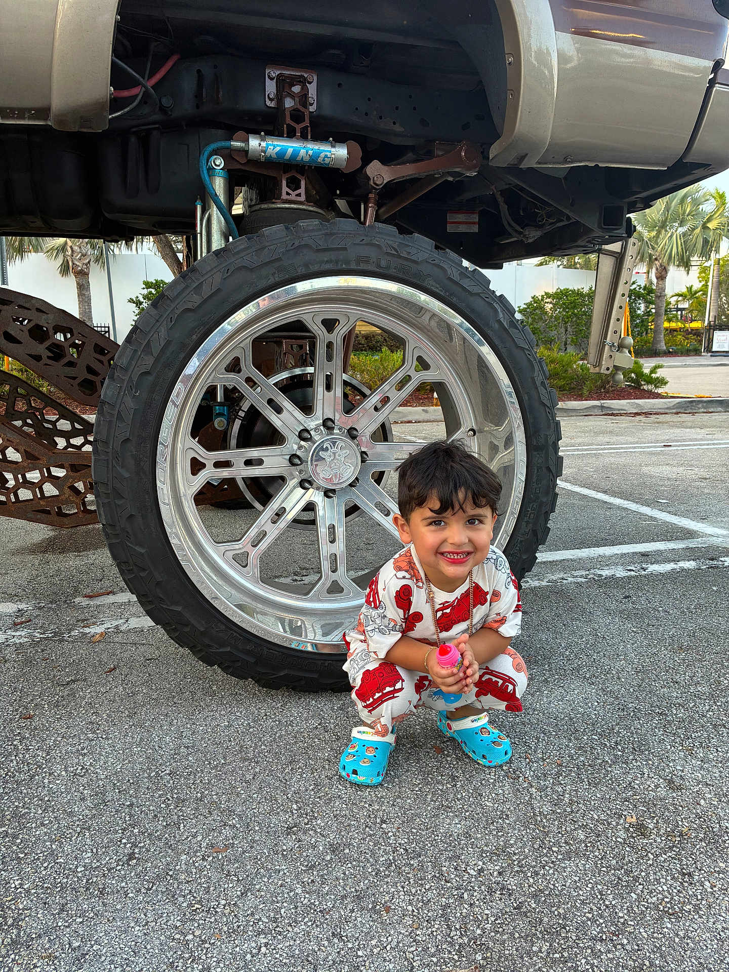 Cyphyr Pineda joined the competition — help win amazing prizes! accessory, asphalt, blue_crocs, boy, casual, child, clothing, cute, daylight, happy, outdoor, parking_lot, playful, smiling, squatting, tire, toy, truck, vehicle, wheel