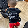 toddler, child, back_view, black_tshirt, red_shorts, sneakers, stroller, indoor, wood_floor, haircut, person, standing, casual_clothing, flooring, footwear, young_child, baby, small_child, clothing, interior