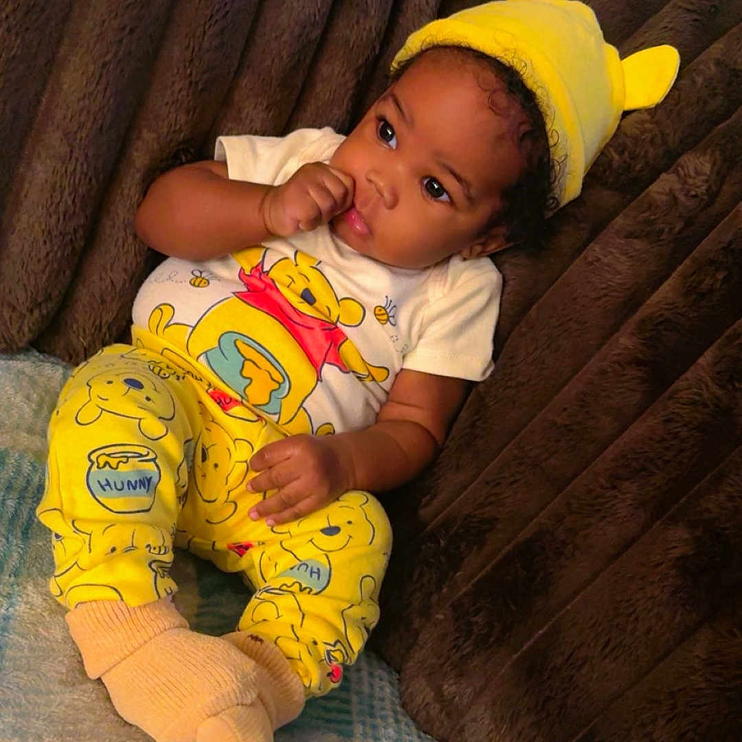 Zahmir joined the competition — help win amazing prizes! baby, child, infant, portrait, smiling, sitting, lying_down, yellow_hat, animal_hat, pajamas, winnie_the_pooh, cartoon, blanket, couch, plush, indoor, cozy, face, hand, sock