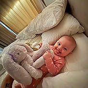 Cassandra joined the competition — help win amazing prizes! baby, bed, pillow, plush_toy, bunny, onesie, smiling, infant, blanket, furniture, indoor, curtains, soft_toy, child, cute, happy, person, cozy, sleepwear, nursery