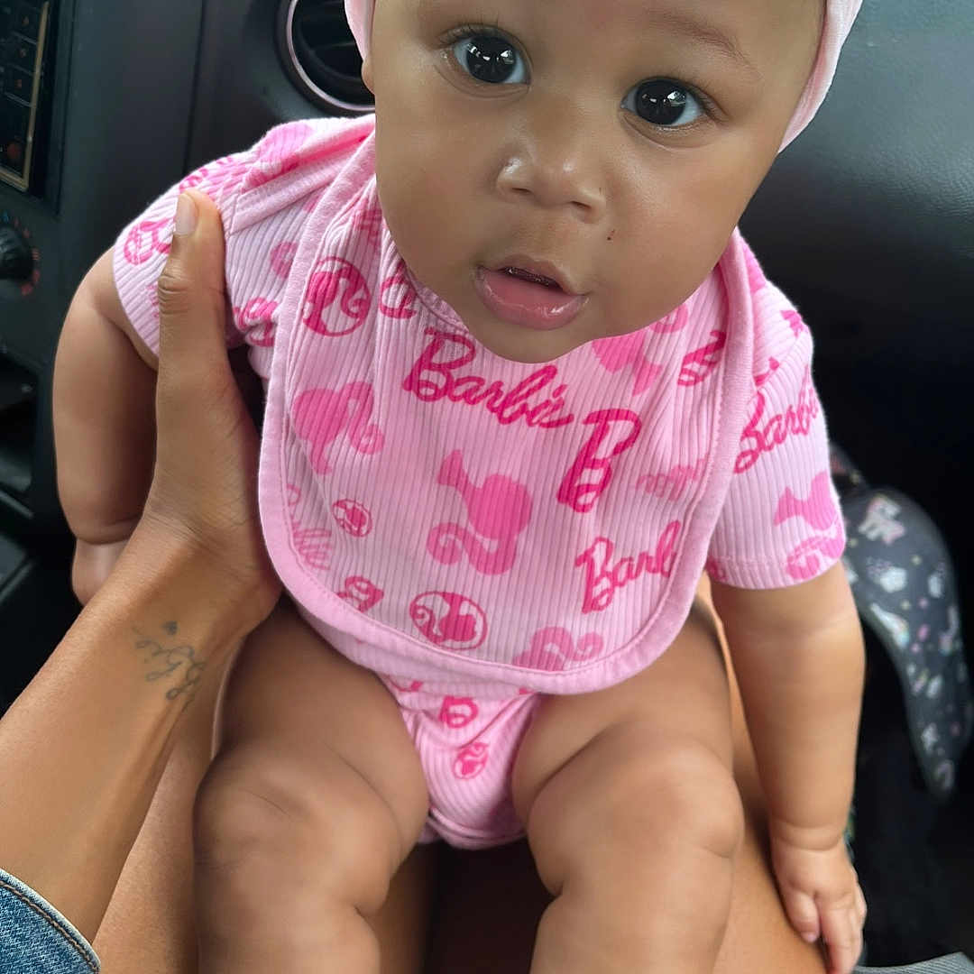 Anaee is registered to the contest to win money with this photo: adorable, baby, bib, car_interior, casual, child, closeup, cute, hand, headwrap, human, infant, legs, looking_at_camera, person, pink_clothing, portrait, seat, sitting, sock