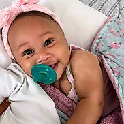 Anaee is registered to the contest to win money with this photo: baby, pacifier, pink_headband, blanket, smiling, infant, face, hand, bed, pillow, cozy, child, cute, sleepwear, portrait, lying_down, comfort, soft, happy, indoors