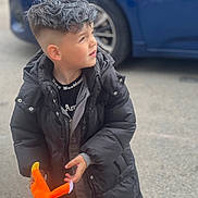 Matthias is registered to the contest to win money with this photo: child, boy, black_jacket, plush_toy, orange_toy, short_hair, silver_hair, car, outdoor, pavement, curious, looking_away, casual_clothing, young_child, street, winter_coat, toy, person, daylight, side_view