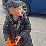 child, boy, black_jacket, plush_toy, orange_toy, short_hair, silver_hair, car, outdoor, pavement, curious, looking_away, casual_clothing, young_child, street, winter_coat, toy, person, daylight, side_view