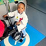 toddler, child, tricycle, bus, wheelchair_symbol, blue_floor, jacket, sneakers, smiling, indoor, public_transport, seat, patterned_seat, handrail, window, person, disability_sign, floor_marking, toy, wheel