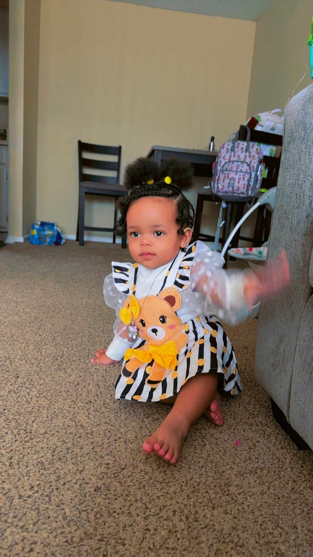 Milani is registered to the contest to win money with this photo: toddler, child, indoor, carpet, dress, teddy_bear, striped_clothing, sitting, waving, hand_motion, hair_buns, necklace, furniture, chair, backpack, floor, room, cute, person, baby