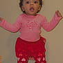 toddler, child, standing, pink_shirt, red_pants, heart_pattern, carpet, wall, curly_hair, pigtails, expression, indoors, person, clothing, footwear, cute, young_child, baby, portrait, casual