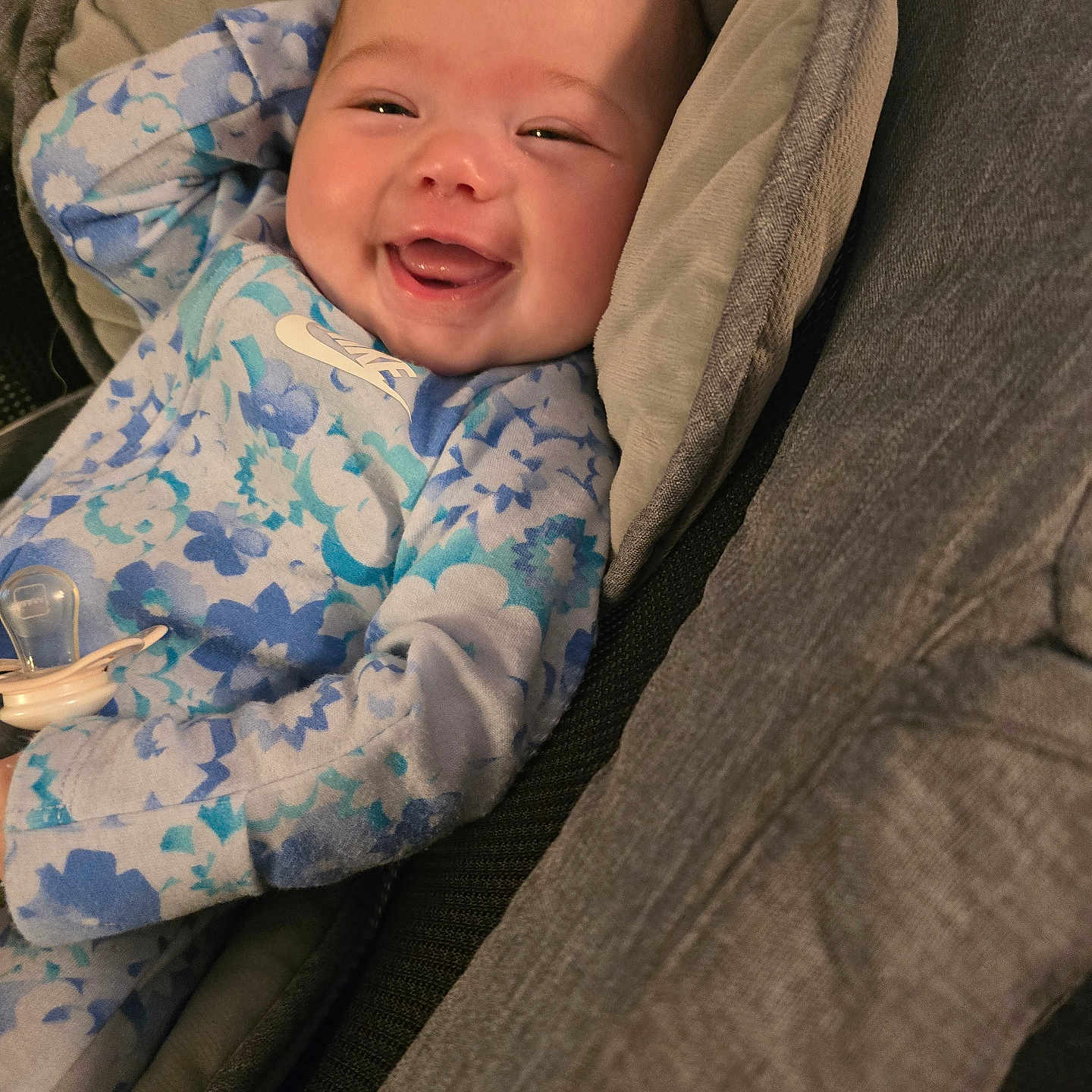 Amelia is registered to the contest to win money with this photo: baby, blue_onesie, child, clothing, comfort, cushion, cute, expression, face, floral_pattern, happy, indoor, infant, joyful, pacifier, portrait, relaxed, seat, smiling, young