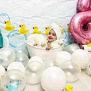 Violet is registered to the contest to win money with this photo: baby, towel, white_tub, balloons, rubber_duck, pink_balloon, number_8, child, smiling, celebration, indoor, decor, festive, cute, white, blue, yellow, background, happy, portrait