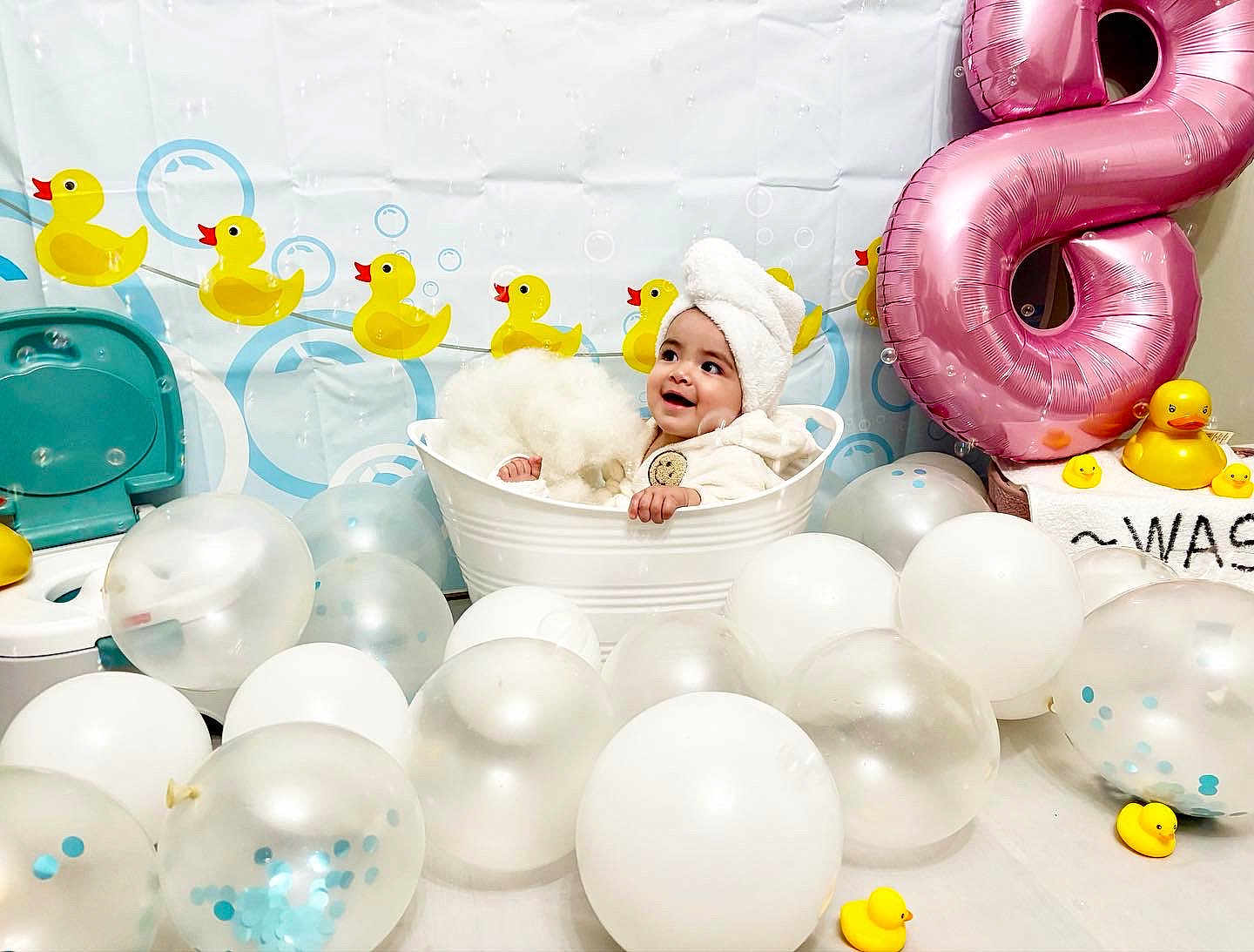 Violet is registered to the contest to win money with this photo: baby, towel, white_tub, balloons, rubber_duck, pink_balloon, number_8, child, smiling, celebration, indoor, decor, festive, cute, white, blue, yellow, background, happy, portrait