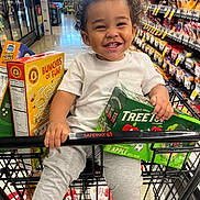 Chance is registered to the contest to win money with this photo: toddler, child, smiling, happy, shopping_cart, grocery_store, supermarket, cereal, snacks, applesauce, food, packaged_goods, shopping, indoors, portrait, curly_hair, white_shirt, gray_pants, cart_handle, safeway