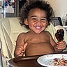 brown_skin, chicken_drumstick, child, curly_hair, dinner, family_meal, food, happy, high_chair, home, indoor, kitchen, messy_eating, plate, portrait, refrigerator, seated, smiling, toddler, tray