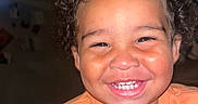 Chance is registered to the contest to win money with this photo: brown_skin, casual_clothing, child, close_up, curly_hair, eyes, face, flash_photography, happy, headshot, home, indoors, joyful, orange_shirt, person, photograph, portrait, smiling, teeth, toddler