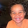 Chance is registered to the contest to win money with this photo: child, toddler, smiling, happy, portrait, curly_hair, brown_skin, face, teeth, eyes, orange_shirt, indoors, flash_photography, close_up, headshot, person, joyful, casual_clothing, home, photograph