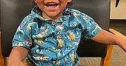 Chance is registered to the contest to win money with this photo: child, toddler, smiling, happy, portrait, sitting, chair, indoor, computer, monitors, curly_hair, dinosaur_shirt, blue_shirt, yellow_shorts, black_shoe, open_mouth, teeth, home, furniture, photography