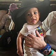 Jakari is registered to the contest to win money with this photo: baby, child, hat, oversized_hat, black_hat, person, hand, bracelet, tattoo, couch, indoor, curious, playful, tongue_out, shirt, shorts, seated, light, window, adult