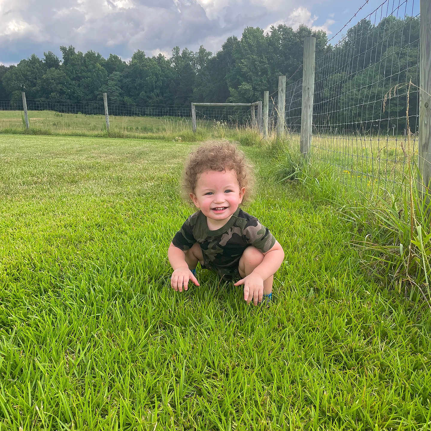 Josiah is registered to the contest to win money with this photo: camouflage_clothing, casual_clothing, child, cloudy_sky, curly_hair, daylight, fence, field, grass, greenery, happy, nature, outdoor, person, playful, rural, smiling, squatting, toddler, young_child