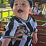 baby, child, smiling, happy, cap, hat, jersey, black_and_white, striped_clothing, outdoor, bench, hand, person, adult, blurred_background, daylight, greenery, fence, portrait, casual_clothing