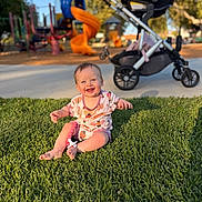 Hazel is registered to the contest to win money with this photo: baby, grass, playground, stroller, smile, sunlight, park, child, outdoor, happy, toy, infant, daylight, nature, greenery, casual_clothing, sitting, cheerful, recreation, family