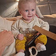 Léo participe au concours pour gagner de l'argent avec cette photo : baby, child, curly_hair, smiling, sitting, remote_control, toy, giraffe_toy, yellow_pants, sweater, lion_king, socks, couch, living_room, lamp, hand, blanket, cozy, indoor, person