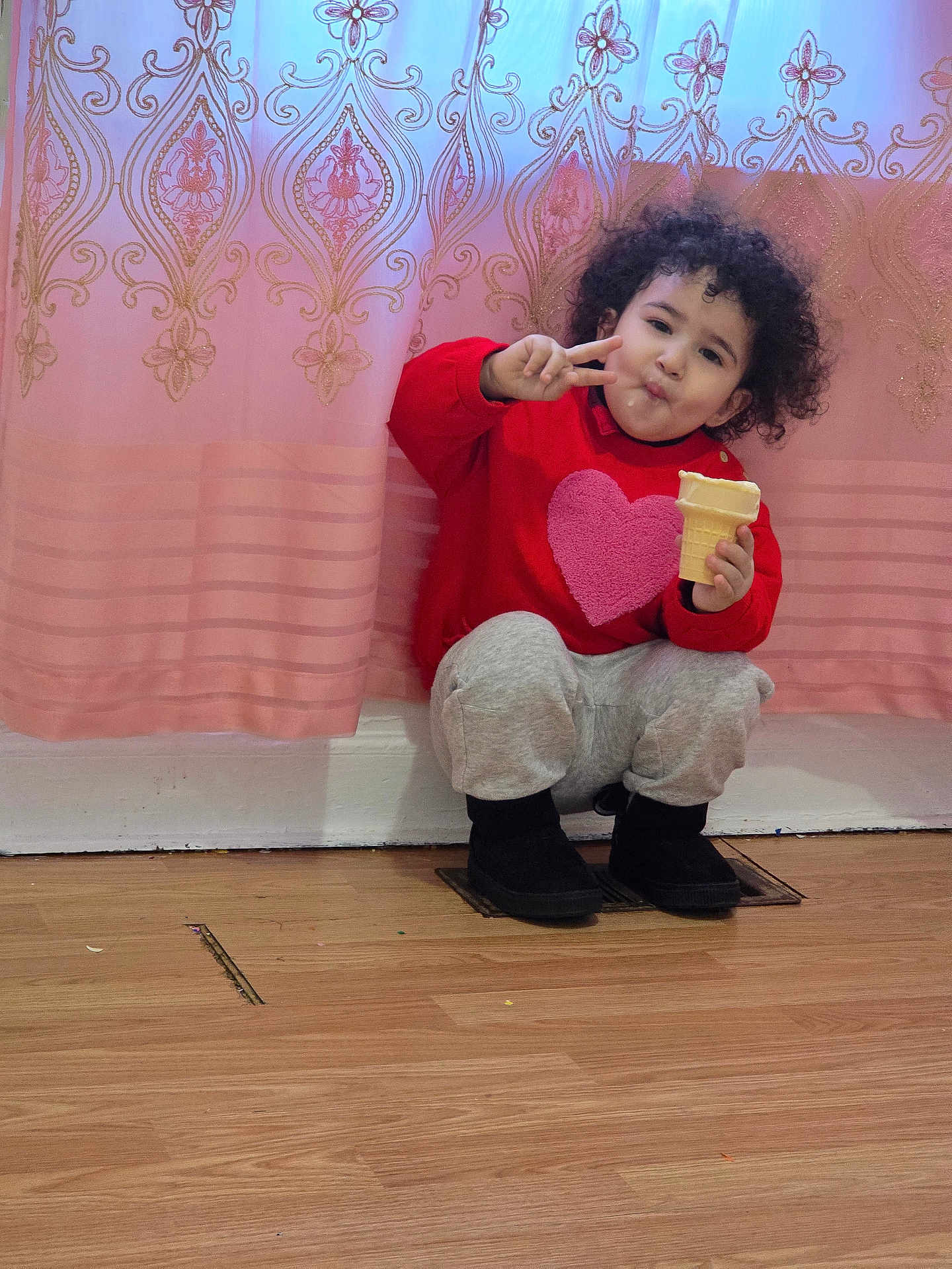 Amer is registered to the contest to win money with this photo: child, toddler, curly_hair, red_sweater, heart_design, ice_cream_cone, peace_sign, pink_curtain, golden_pattern, wooden_floor, black_boots, gray_pants, indoor, cute, happy, portrait, sitting, playful, smiling, cozy