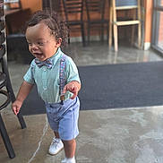 Timothy is registered to the contest to win money with this photo: toddler, child, standing, walking, smiling, bow_tie, suspenders, shorts, shirt, dress_shoes, white_shoes, curly_hair, portrait, indoor, cafe, restaurant, chairs, chalkboard, window, concrete_floor