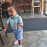 toddler, child, standing, walking, smiling, bow_tie, suspenders, shorts, shirt, dress_shoes, white_shoes, curly_hair, portrait, indoor, cafe, restaurant, chairs, chalkboard, window, concrete_floor