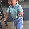 Timothy is registered to the contest to win money with this photo: toddler, child, boy, portrait, standing, walking, smiling, bow_tie, suspenders, shirt, shorts, dress_shoes, curly_hair, outdoor, restaurant, cafe, chairs, concrete_floor, fashion, happy