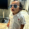 child, toddler, portrait, sunglasses, standing, kitchen, indoors, white_shirt, button_up_shirt, shorts, curly_hair, open_mouth, barstool, sink, cabinets, tile_floor, home, fashion, posing, smiling
