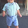 Timothy is registered to the contest to win money with this photo: toddler, child, boy, portrait, standing, walking, bow_tie, suspenders, shorts, shirt, sneakers, curly_hair, smiling, outdoor, entryway, chairs, concrete, fashion, cute, full_body