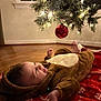 baby, child, christmas_tree, ornament, red_ball, blanket, brown_costume, floor, happy, smiling, holiday, festive, indoor, wooden_floor, lights, pine_tree, cute, infant, cozy, celebration