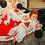 baby, infant, christmas_tree, christmas_ornaments, plush_toy, pacifier, holiday, festive_clothing, red_bow, white_pajamas, floor, decorations, indoor, carpet, cute, child, holiday_season, celebration, toy, relaxing