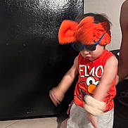 Zaire is registered to the contest to win money with this photo: child, toddler, elmo, sunglasses, headband, orange, red, tank_top, shorts, indoor, tile_floor, person, motion_blur, black_door, playful, fun, accessories, cute, expression, home