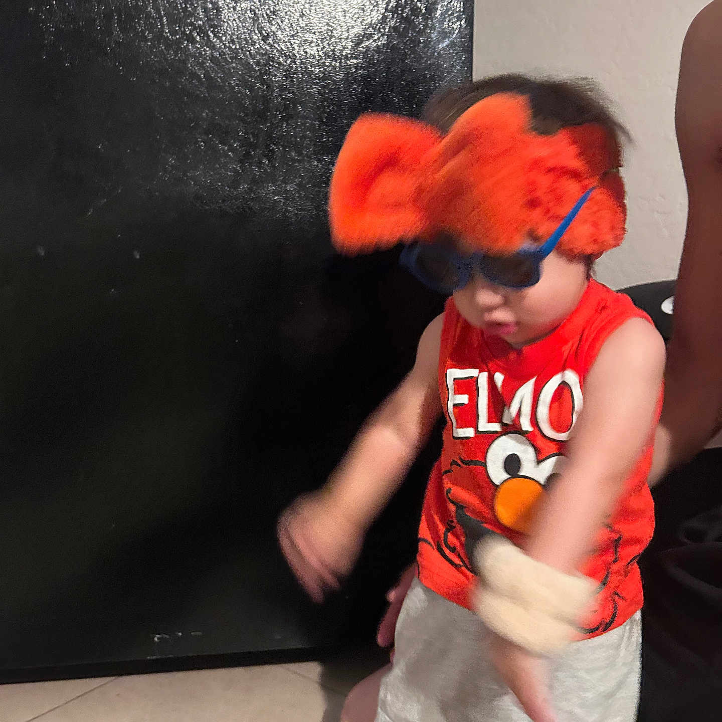 Zaire is registered to the contest to win money with this photo: child, toddler, elmo, sunglasses, headband, orange, red, tank_top, shorts, indoor, tile_floor, person, motion_blur, black_door, playful, fun, accessories, cute, expression, home