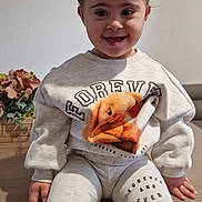 Isla is registered to the contest to win money with this photo: toddler, child, smile, sitting, sweatshirt, leggings, hair_buns, portrait, indoor, table, flower_arrangement, framed_photos, cute, happy, clothing, young_child, family, casual, cozy, home